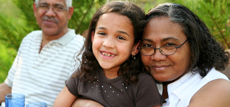 a smiling girl sitting on a woman's lap with the woman hugging her and smiling with a man smiling in the background